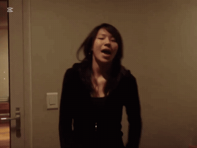 A Woman In A Black Jacket Stands In Front Of A Wall With An X In The Corner GIF