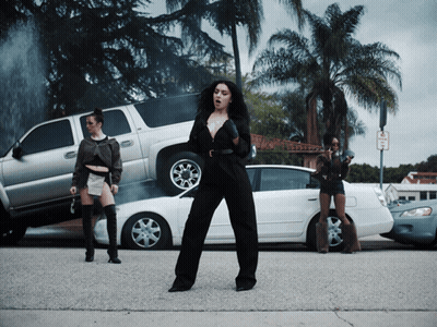 A Woman In A Black Jumpsuit Is Standing In Front Of A Stack Of Cars And A No Parking Sign GIF