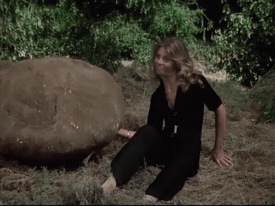 A Woman In A Black Jumpsuit Sits In The Grass Next To A Large Rock GIF