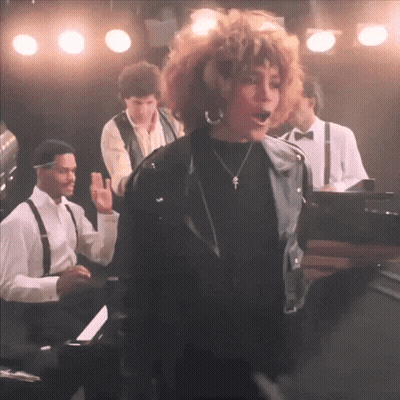 A Woman In A Black Leather Jacket Is Singing On Stage GIF