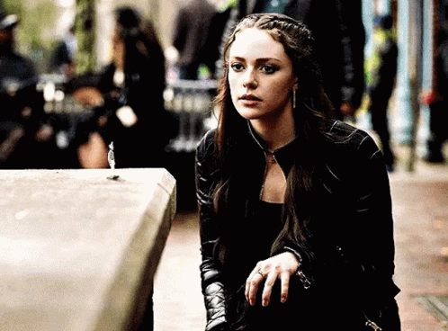 A Woman In A Black Leather Jacket Is Sitting On A Sidewalk Looking At Something . GIF