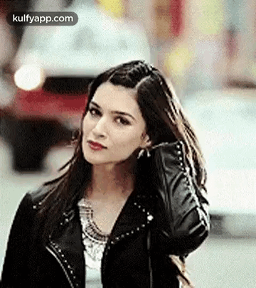 A Woman In A Black Leather Jacket Is Standing On A Street Holding Her Hair . GIF