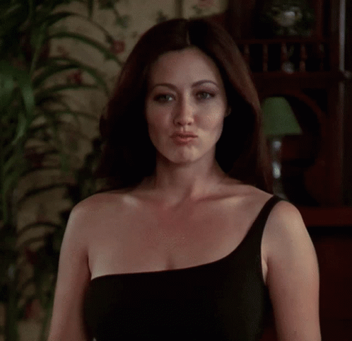 A Woman In A Black One Shoulder Dress Is Standing In Front Of A Lamp . GIF
