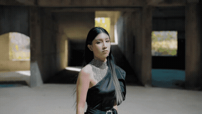 A Woman In A Black One Shoulder Dress Stands In A Dark Room GIF