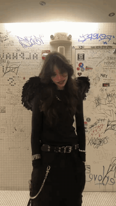 A Woman In A Black Outfit Is Standing In Front Of A Wall That Has Graffiti On It Including The Word Ovoks GIF