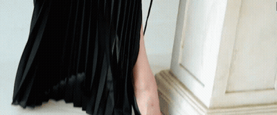 A Woman In A Black Pleated Skirt Is Standing Next To A White Column GIF