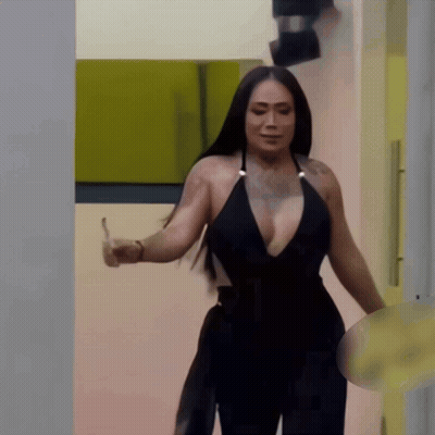 A Woman In A Black Plunging Neckline Top Is Standing In A Hallway GIF
