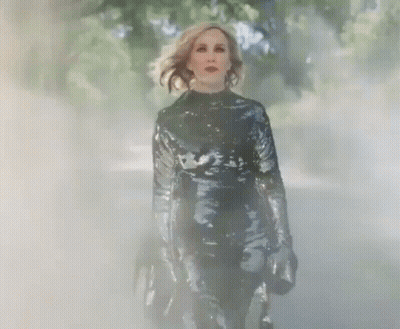 A Woman In A Black Sequined Jumpsuit Is Walking Through A Foggy Forest . GIF