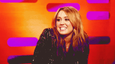 A Woman In A Black Sequined Top Smiles In Front Of A Purple And Orange Background GIF