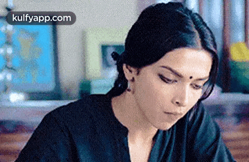 A Woman In A Black Shirt Is Sitting At A Table Looking Down At Something . GIF