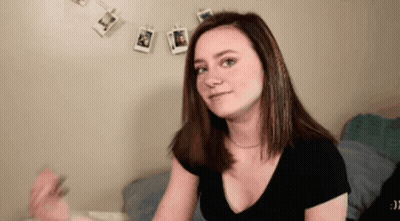 A Woman In A Black Shirt Is Sitting On A Bed In Front Of A Wall With Pictures Hanging On It . GIF