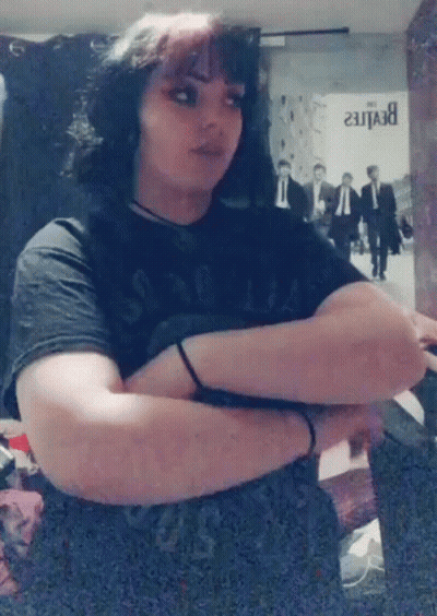 A Woman In A Black Shirt Is Standing With Her Arms Crossed In Front Of A Beatles Poster . GIF