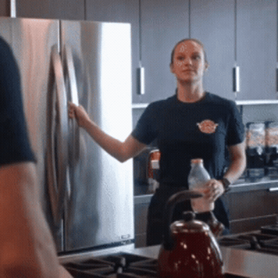 A Woman In A Black Shirt Stands In A Kitchen Holding A Bottle GIF