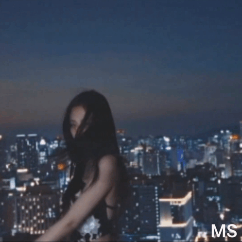 A Woman In A Black Strapless Dress Is Standing In Front Of A City Skyline At Night . GIF