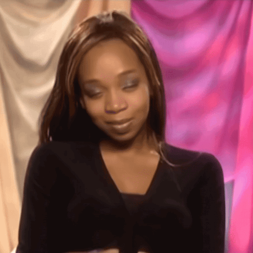 A Woman In A Black Sweater Is Smiling In Front Of A Pink Curtain .. GIF