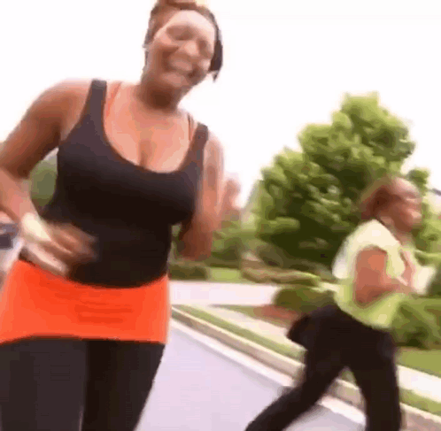 A Woman In A Black Tank Top And Orange Skirt Is Jogging Down A Street . GIF