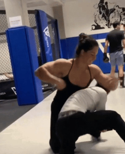 A Woman In A Black Tank Top Holds A Man In A Gym GIF