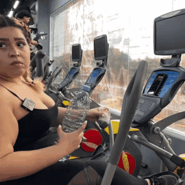 A Woman In A Black Tank Top Is Sitting On An Elliptical Machine Holding A Bottle Of Water GIF