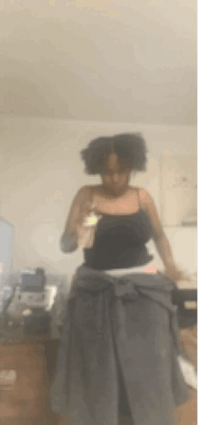 A Woman In A Black Tank Top Is Standing In A Living Room Holding Her Hair . GIF