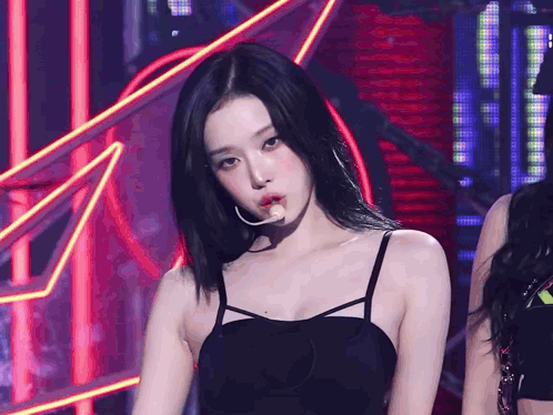 A Woman In A Black Tank Top With A Microphone In Her Mouth Looks At The Camera GIF