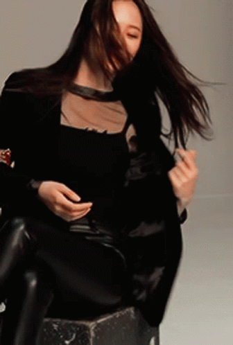 A Woman In A Black Top And Black Pants Is Sitting On A Stool With Her Hair Blowing In The Wind GIF