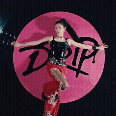 A Woman In A Black Top And Red Pants Stands In Front Of A Pink Circle That Says Drip GIF