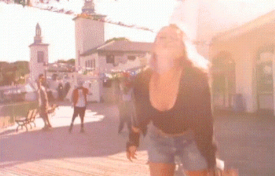 A Woman In A Black Top And Shorts Is Standing In Front Of A Crowd Of People In A Park . GIF
