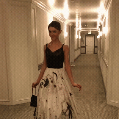 A Woman In A Black Top And White Skirt Is Walking Down A Hallway GIF