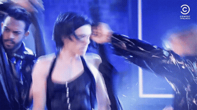 A Woman In A Black Top Is Dancing With A Man In A Black Jacket Behind Her .. GIF