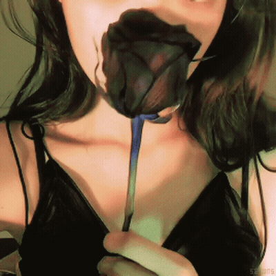 A Woman In A Black Top Is Holding A Black Rose In Her Mouth GIF