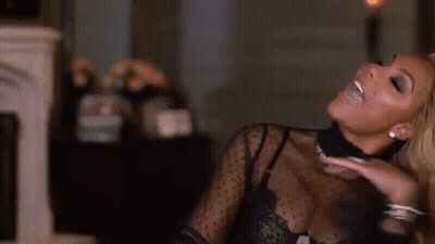 A Woman In A Black Top Is Smiling And Sitting In A Living Room . GIF