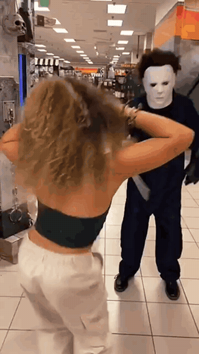 A Woman In A Black Top Is Standing Next To A Man In A Mask In A Store . GIF