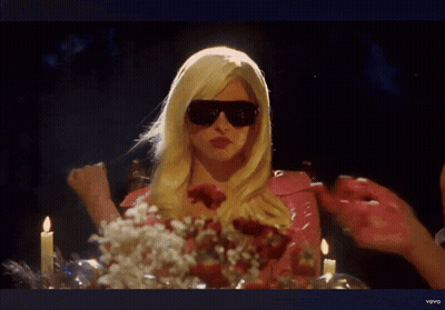 A Woman In A Blonde Wig And Sunglasses Is Sitting At A Table With Candles And Flowers GIF