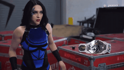 A Woman In A Blue And Black Wrestling Outfit Stands In Front Of A Peacock Logo GIF