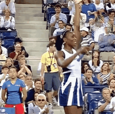 A Woman In A Blue And White Dress Is Raising Her Fist In The Air In Front Of A Crowd GIF