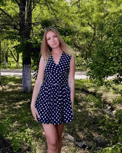 A Woman In A Blue And White Polka Dot Dress Stands In A Park GIF