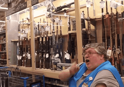 A Woman In A Blue Apron Is Standing In Front Of A Walmart Display Of Guns GIF