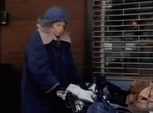 A Woman In A Blue Coat Is Pushing A Shopping Cart With A Bag Of Trash . GIF