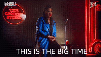 A Woman In A Blue Coat Is Sitting In Front Of A Neon Sign That Says The Comedy Store GIF