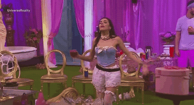A Woman In A Blue Corset Is Dancing In A Room With Purple Curtains And Gold Chairs . GIF
