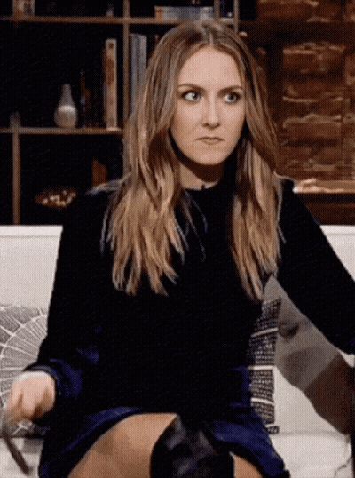 A Woman In A Blue Dress And Black Boots Is Sitting On A Couch Making A Funny Face . GIF