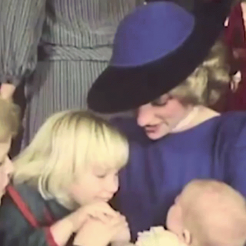 A Woman In A Blue Dress And Black Hat Is Holding A Baby In Her Arms . GIF