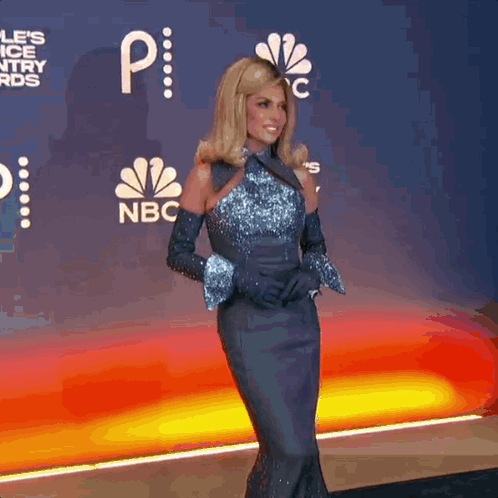 A Woman In A Blue Dress And Gloves Is Standing On A Red Carpet With A Nbc Logo In The Background GIF