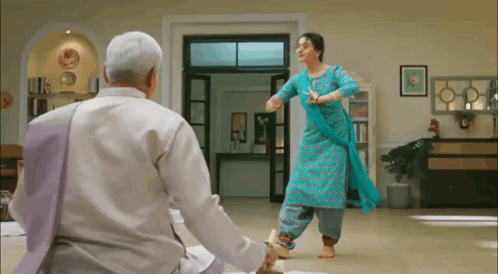 A Woman In A Blue Dress Is Dancing In A Living Room While A Man Looks On . GIF