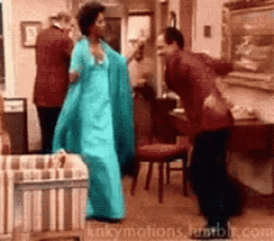 A Woman In A Blue Dress Is Dancing With A Man In A Red Suit In A Living Room GIF