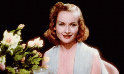 A Woman In A Blue Dress Is Smiling In Front Of A Vase Of Flowers GIF