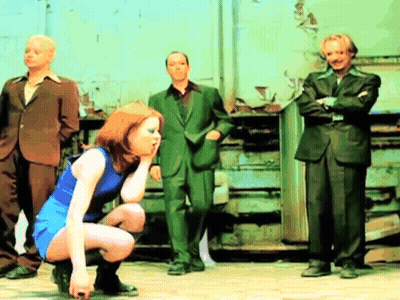 A Woman In A Blue Dress Is Squatting Down In Front Of Three Men In Suits GIF