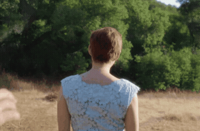 A Woman In A Blue Dress Is Standing In A Field With Trees In The Background GIF