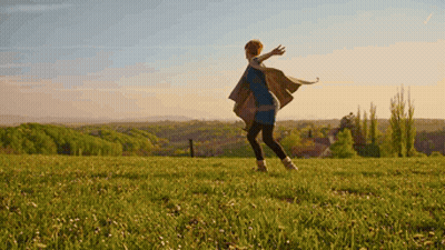 A Woman In A Blue Dress Is Standing In A Grassy Field With Her Arms Outstretched GIF
