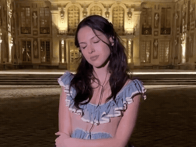 A Woman In A Blue Dress Is Standing In Front Of A Building With Her Eyes Closed GIF